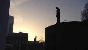 This picture shows a silhouette stood on a tall block on the right of the image, gazing down to the bottom-left which has a lower block. There is funky lighting in the sky. 